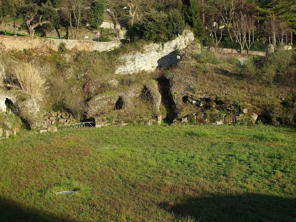 A grassy arena seen from its edge, with a curving bank of rough masonry and dark arched openings set into the slope beyond it, and trees and houses on the rim above.