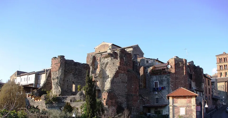 Tall broken walls of brick and rubble rising among the houses of a hillside quarter, with a brick bell tower on the right against a clear sky.