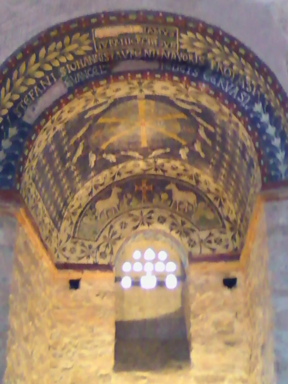 A barrel-vaulted niche covered in mosaic on a deep blue ground, with a large monogram in rings at the centre, small figures of doves and lambs, and a band of lettering round the arch above a small window.