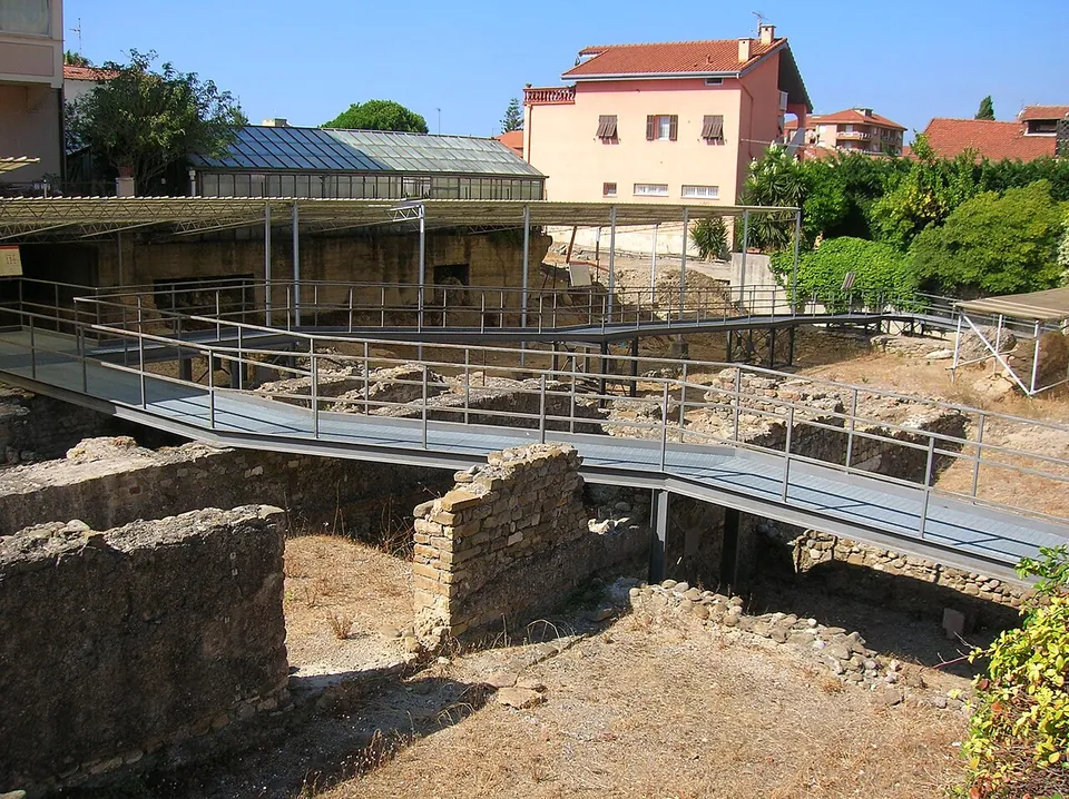 Low ruined walls of rubble masonry in a dry, sandy excavation, crossed by a raised metal walkway with railings, with a glass-roofed building and a pink house behind.