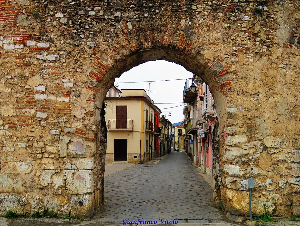 A round-headed arch through a thick wall of rough stone, its arch outlined in red brick, with a narrow paved street of pastel houses running away through the opening.