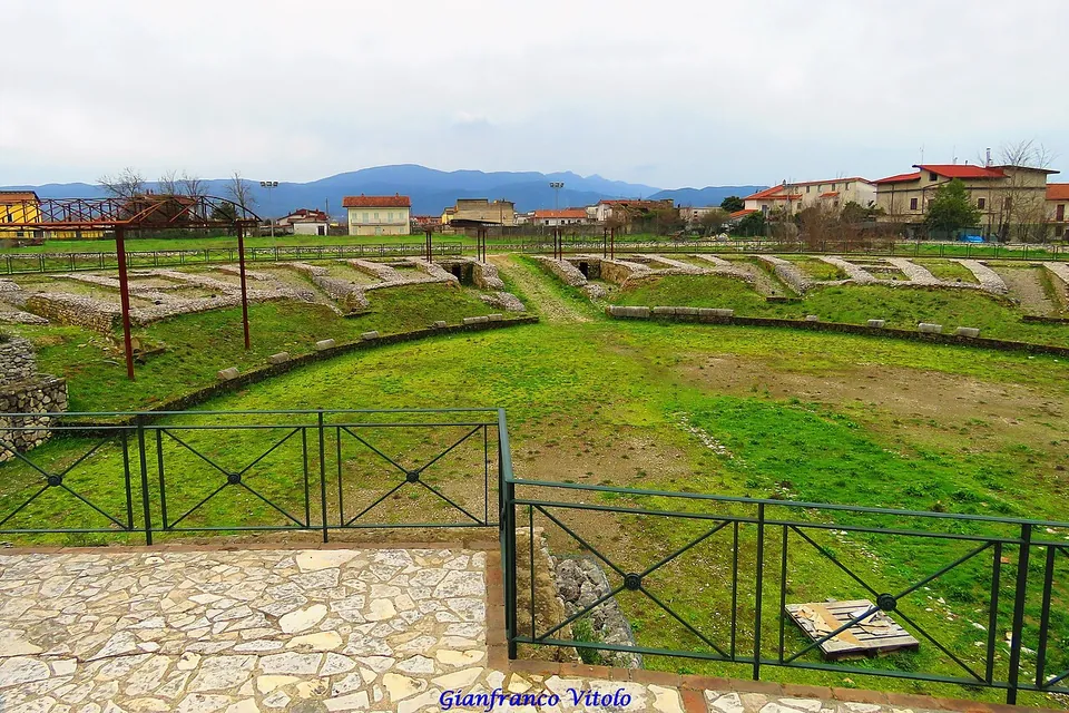 An oval arena of grass inside a low ring of stone walls and grassy banks, seen over a green metal railing from a paved terrace, with houses and mountains behind.