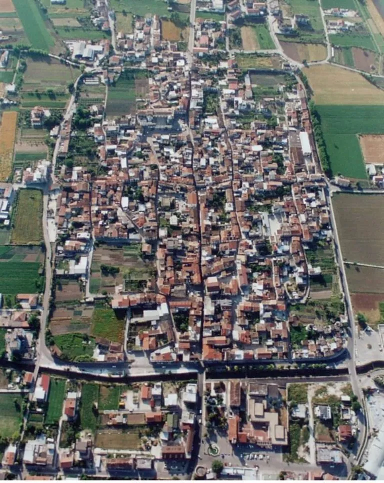 An aerial view looking down on a compact town of red-tiled roofs packed into a rectangle crossed by straight streets, with a wide road along one side and open fields around it.