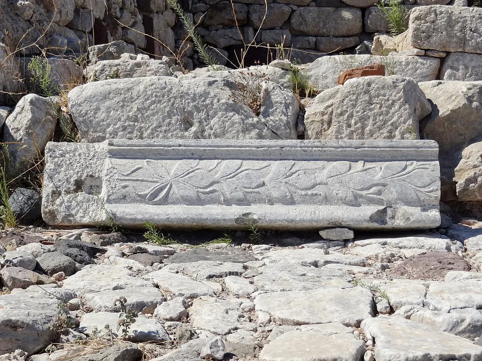 A long marble block carved with a leafy scroll between plain mouldings, lying on a street of rough paving stones in front of a wall of large blocks.