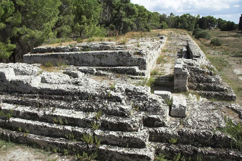 Weathered limestone steps and squared blocks of a long, low platform, a stepped cut rising in the middle distance, with pine trees behind.
