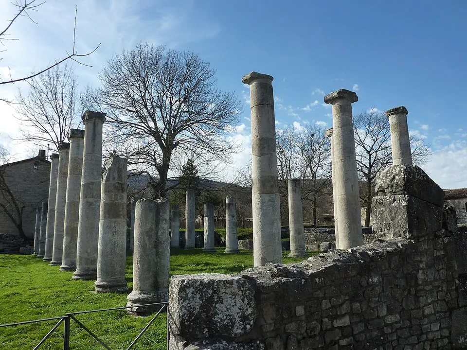 Two rows of plain round stone columns standing in mown grass behind a low rubble wall, some at full height with simple capitals, others broken off part-way, with bare trees and a stone farmhouse behind.