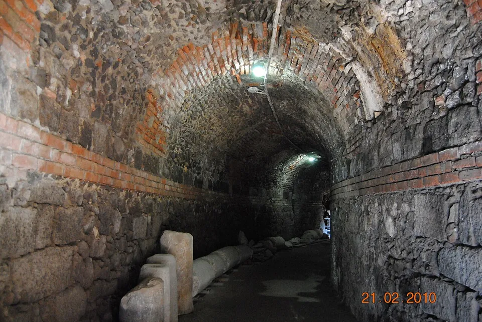 A long, dimly lit vaulted corridor of rough lava stone with bands of brick, lined along its left wall with fragments of stone columns and blocks.