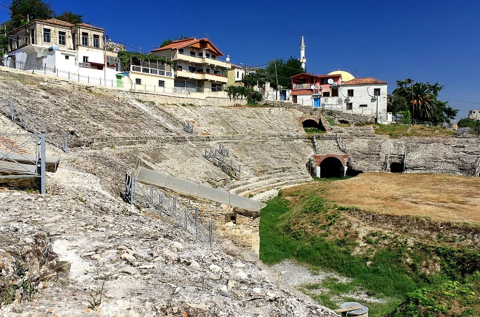 Rows of worn stone seating and ruined walls curving round a grassy arena, with metal railings on the slopes and white houses above the rim.