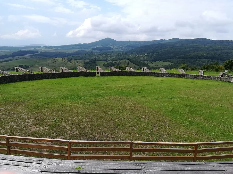 The oval arena of an amphitheatre, a mown lawn enclosed by a low stone wall with pillars, seen over a wooden fence, with forested hills and a wide sky beyond.