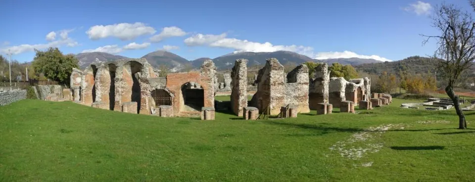 A wide panorama across mown grass to a curving row of ruined brick piers and arches, with a line of low mountains behind.