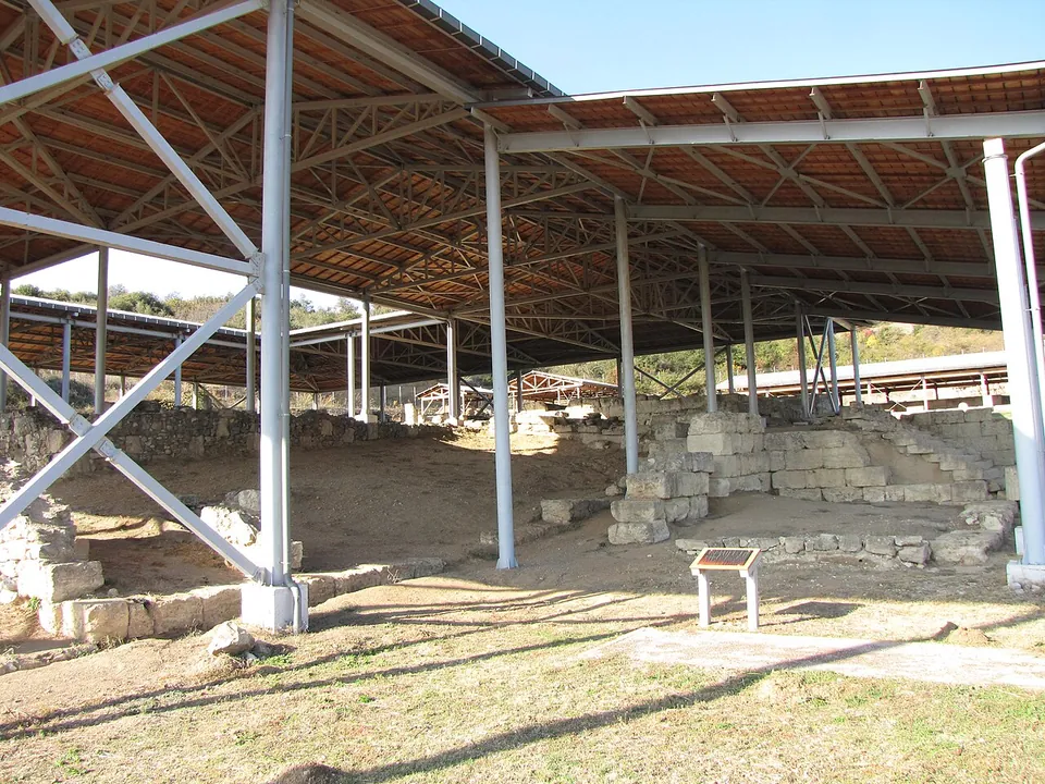 Excavated stone walls, steps and a sunken floor under a long modern steel roof on slender columns, with an information sign in front and wooded hills beyond.
