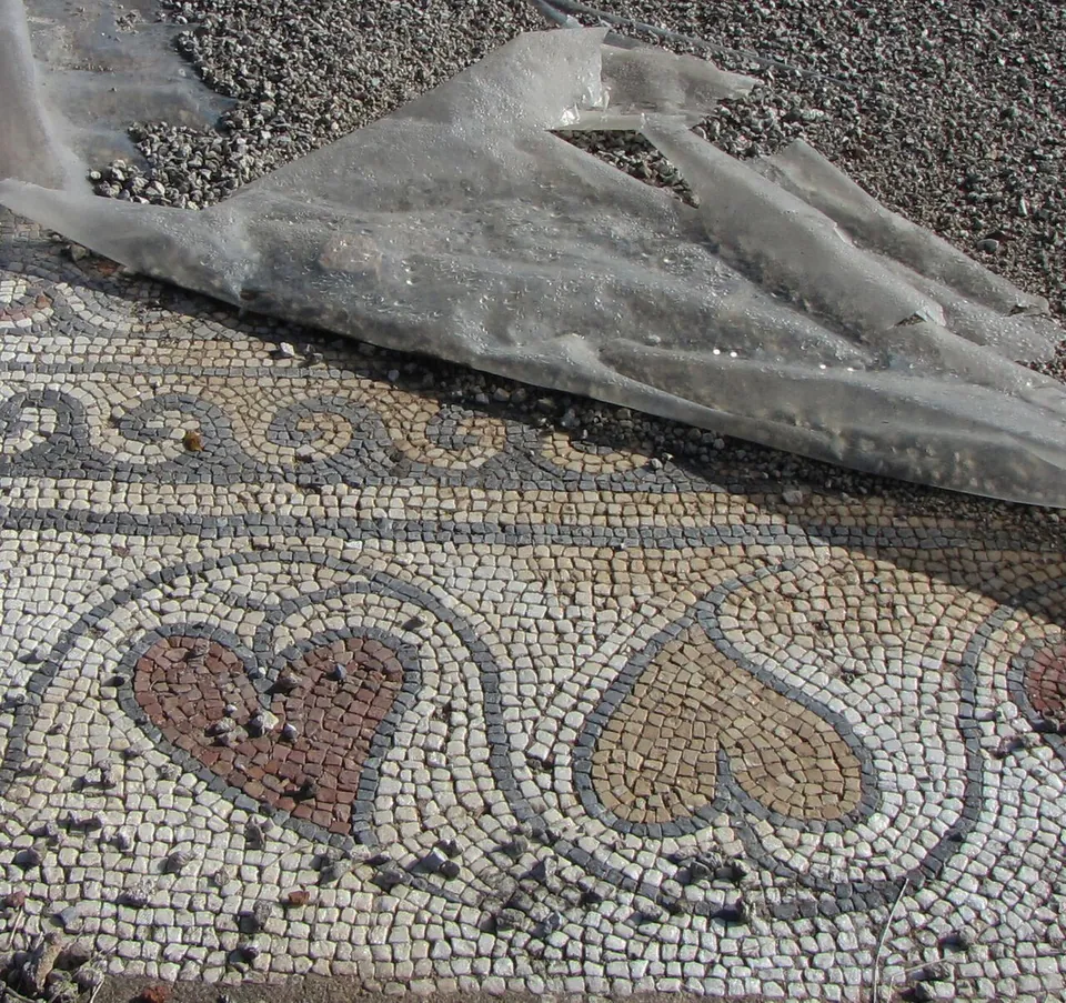A floor mosaic of small cream, grey and red stones with a wave border and a row of heart-shaped motifs, partly covered by a sheet of clear plastic weighed down with gravel.