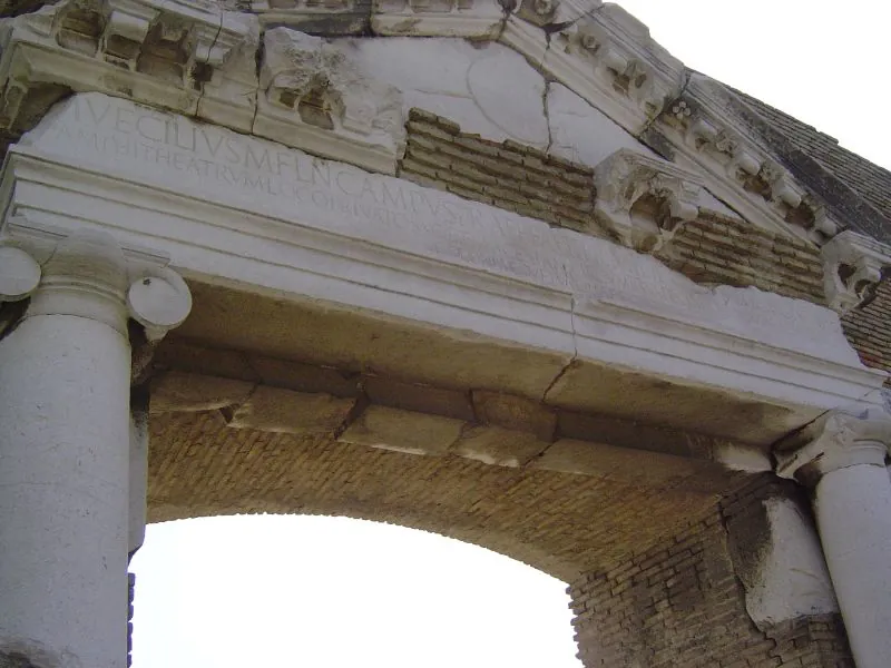 Looking up at a stone lintel resting on Ionic capitals, carved with a Latin inscription in capital letters, under a cornice of carved stone and brick.