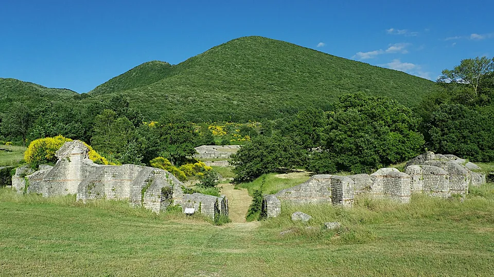 Grey stone walls of the amphitheatre, broken by an opening, standing in mown grass in front of a steep, wooded green hill.