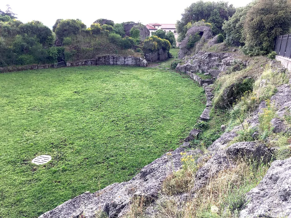 A mown oval of grass enclosed on one side by a rough curving face of grey bedrock, overgrown with plants, with low walls and trees at the far end.