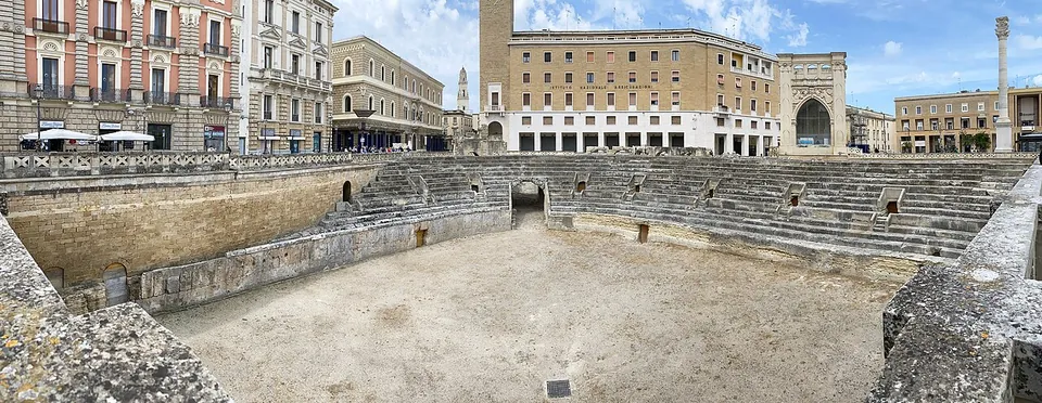 A wide panorama of a sunken oval arena of pale stone, with tiers of stone seating curving round the far side and passages opening beneath them, and town buildings, an arched stone front and a tall column standing round its rim at street level.