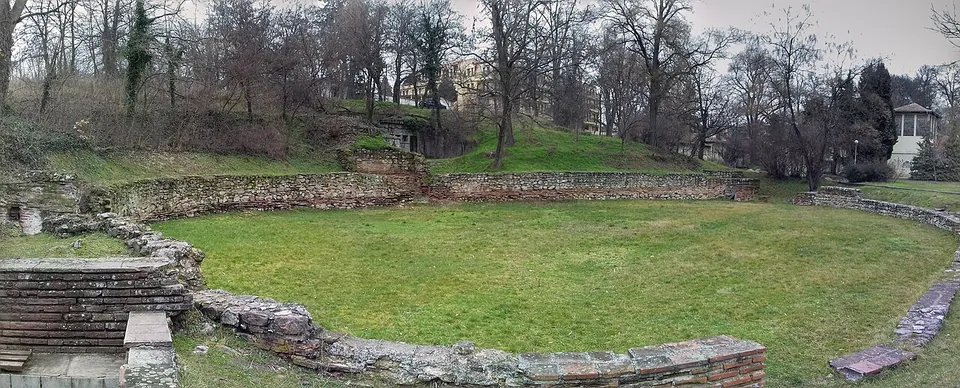 A grassed oval arena edged by a low curving wall of stone, with stone steps on the left and bare winter trees on the slope beyond.