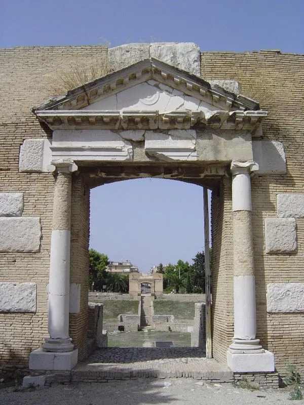 A doorway of pale stone with a column on each side, the shafts patched with newer white stone, and a triangular pediment above, set in a wall of small yellow blocks, looking through across the arena to a second gateway on the far side.