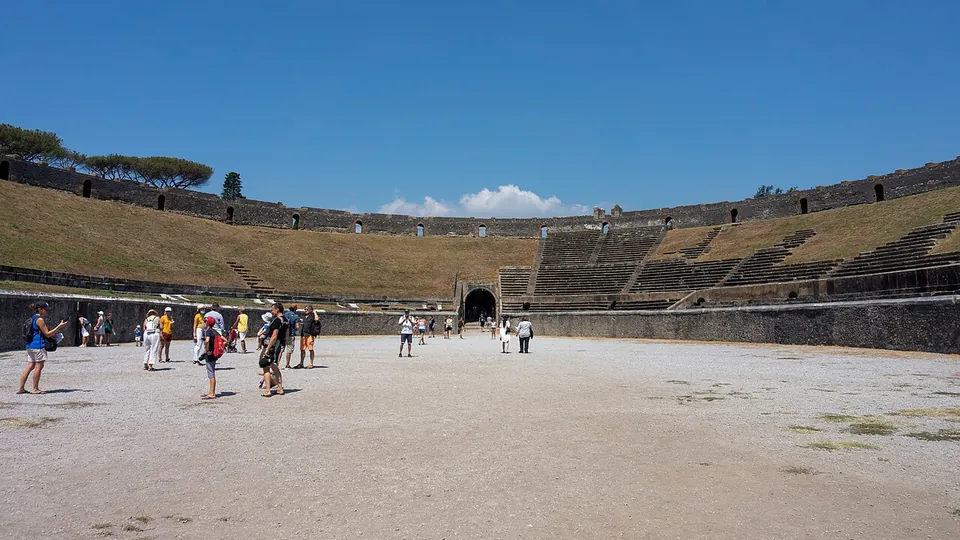 The oval arena of the amphitheatre seen from its sandy floor, visitors walking across it, grassed banks of stone seating rising all round and an arched entrance in the far wall.