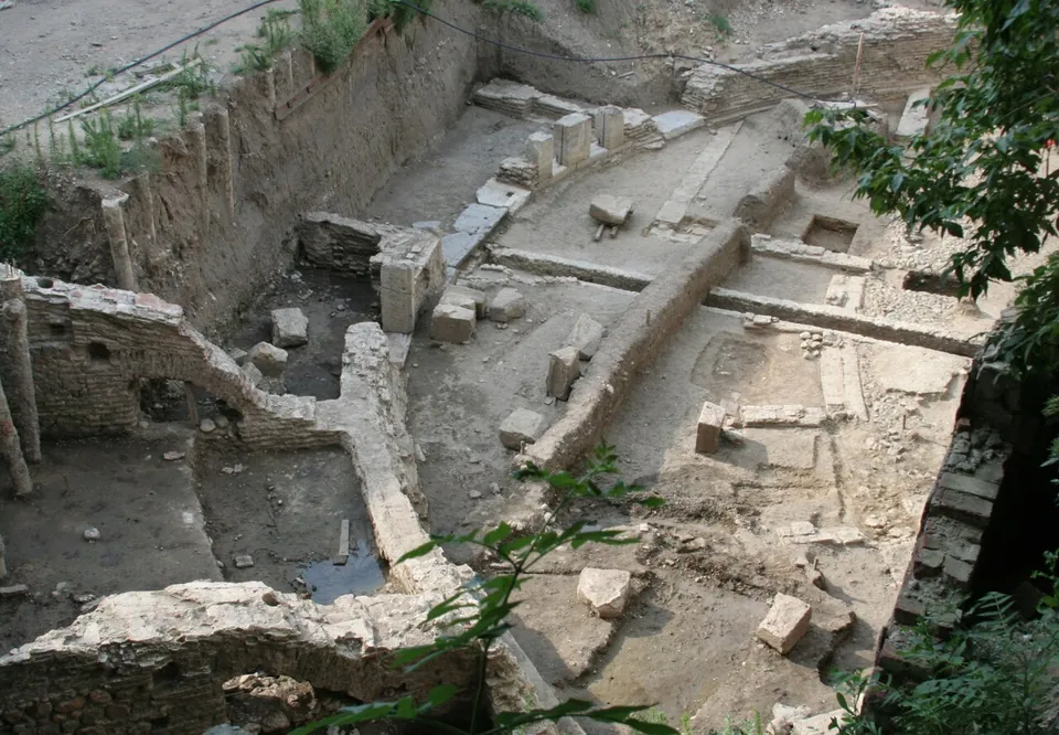An open excavation seen from above, with stone and brick walls, a curving wall line and scattered blocks in a dug-out pit edged by trees and earth banks.