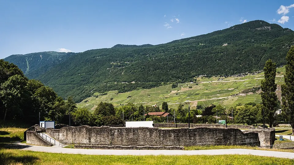 The long, low outer wall of the amphitheatre in rough stone across a lawn, with vineyard terraces and a steep wooded mountain rising behind under a blue sky.