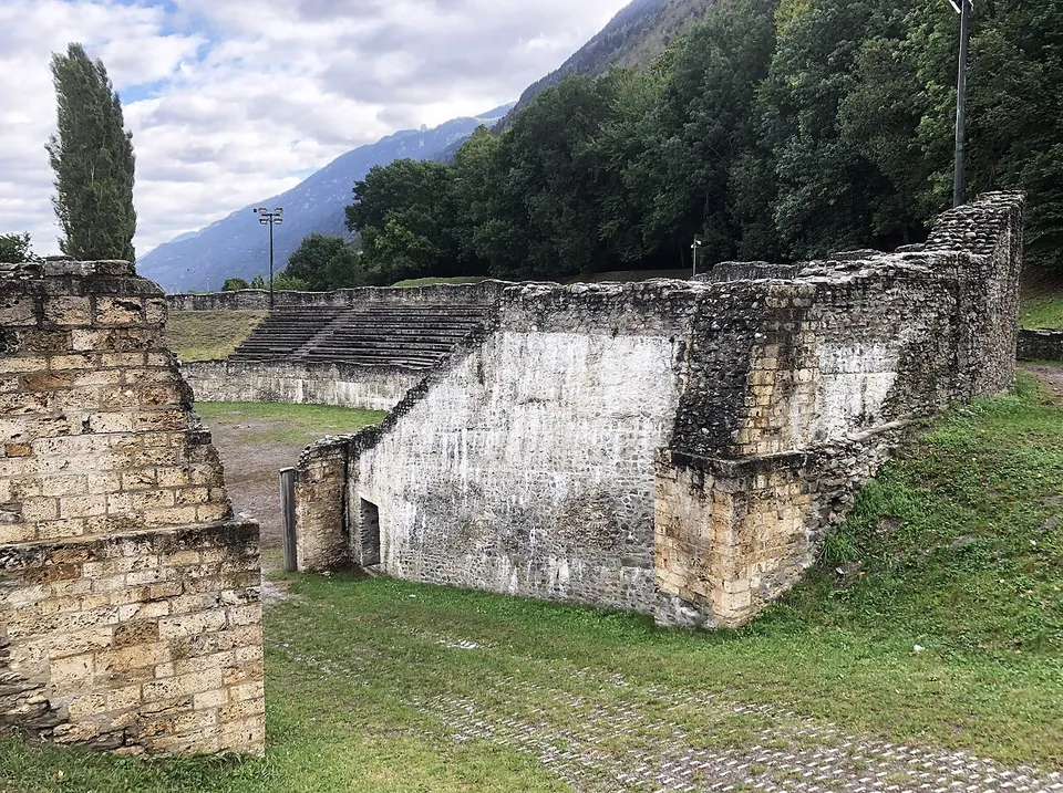 Heavy walls of squared yellowish stone forming an entrance passage into the arena, with grass in front, the seating banks behind and wooded mountains beyond.