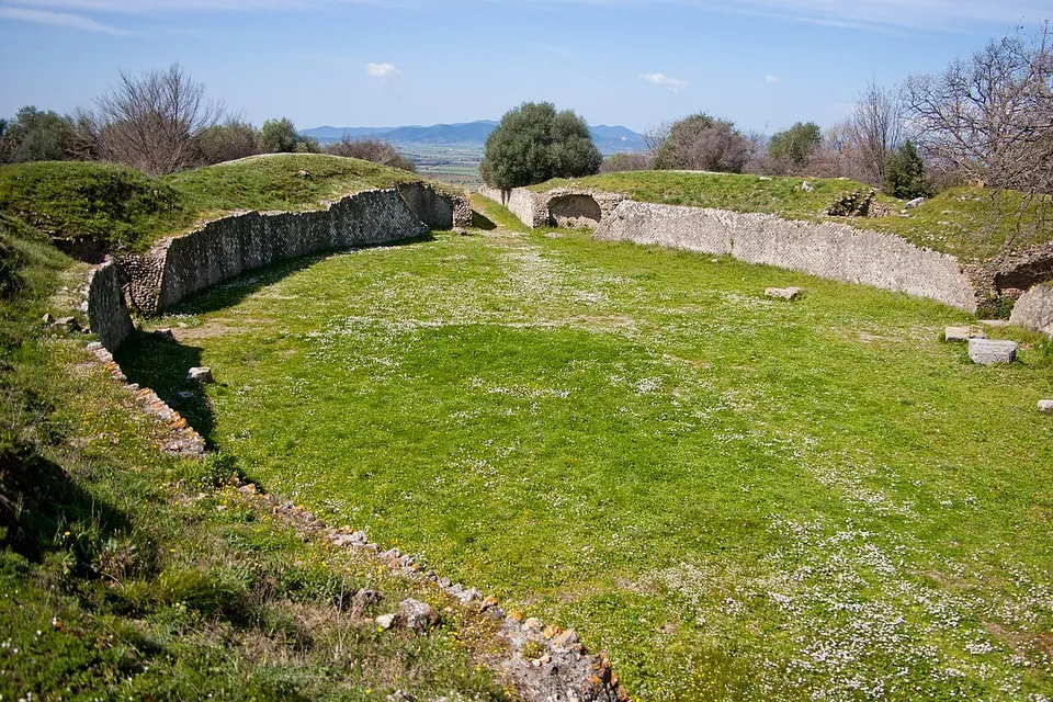 An oval arena of mown grass enclosed by a curving stone retaining wall, with grassy banks above it, an opening in the far wall and a plain and low hills on the horizon.