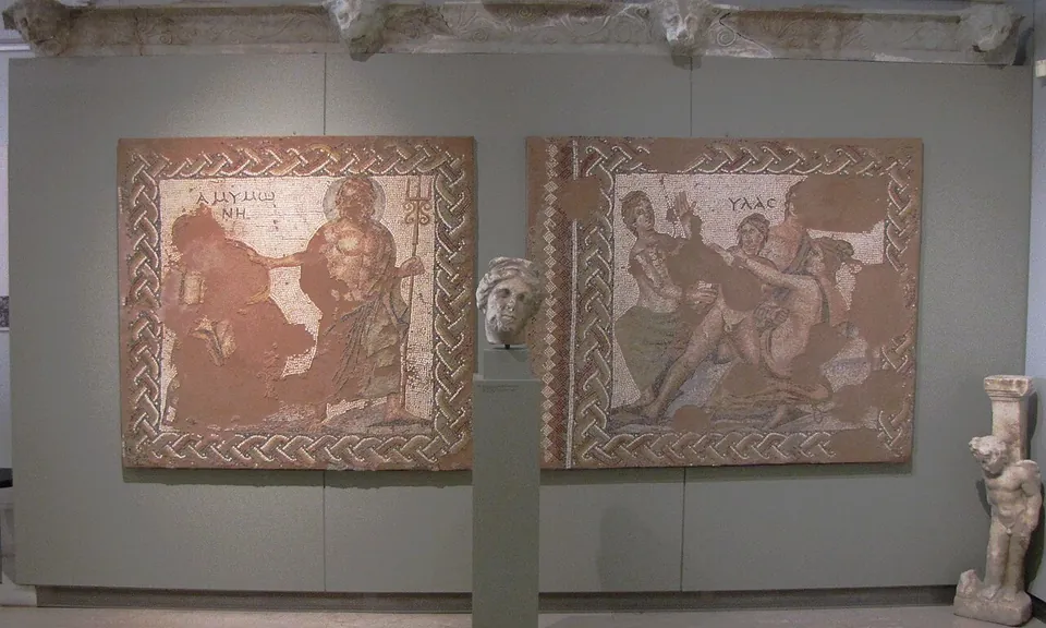 Two square floor mosaics hung side by side on a grey wall, each framed by a plaited border and showing mythological figures with their names in Greek letters, with a marble head on a plinth between them.