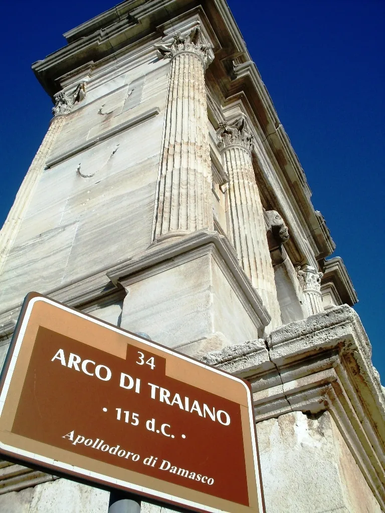 A brown street sign reading "Arco di Traiano, 115 d.C., Apollodoro di Damasco" in front of the arch's marble columns, seen from below against a deep blue sky.