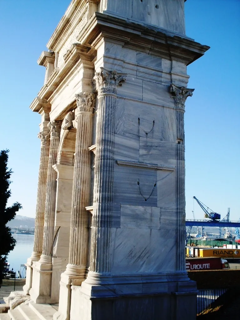 The side of the arch from below, fluted columns with Corinthian capitals against weathered marble blocks pitted with small holes, cranes of the port visible beyond.
