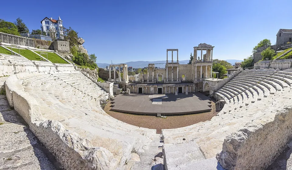 A wide panorama of the whole Roman theatre from the top of the seating, the marble rows curving round a semicircular stage floor towards a two-storey columned stage building, with a white house on the hill to the left and distant mountains under a clear sky.