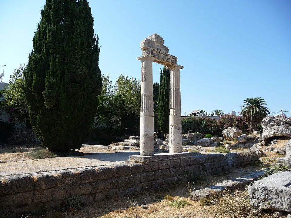 Two fluted marble columns carrying a piece of entablature, standing on a stone wall in the ruins of the agora, beside a tall cypress, under a blue sky.