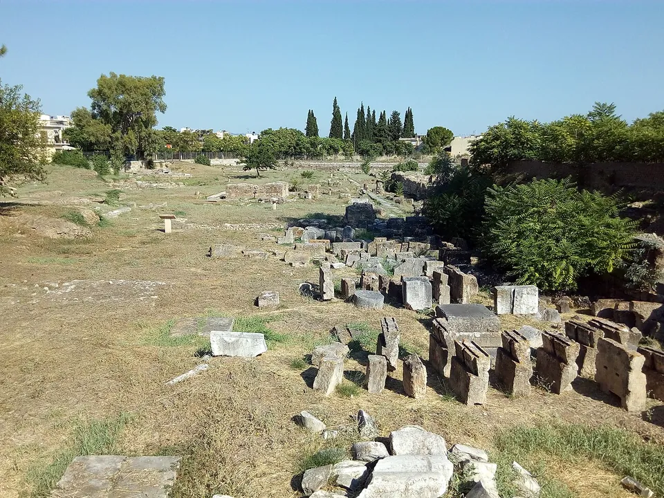 An excavated field strewn with rows of stone blocks and column pieces among dry grass, with cypresses and houses at the far edge.