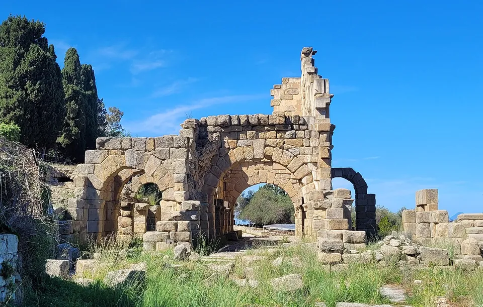 A ruined building of large squared sandstone blocks pierced by wide round arches, open as passages, standing among grass and scattered blocks against a blue sky.