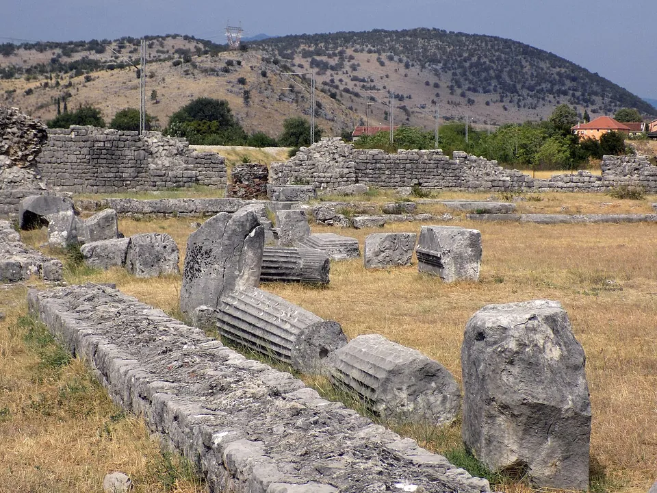 A dry grassy field strewn with fallen column drums and carved stone blocks beside a low line of wall, with ruined walls several metres high and scrubby hills behind.