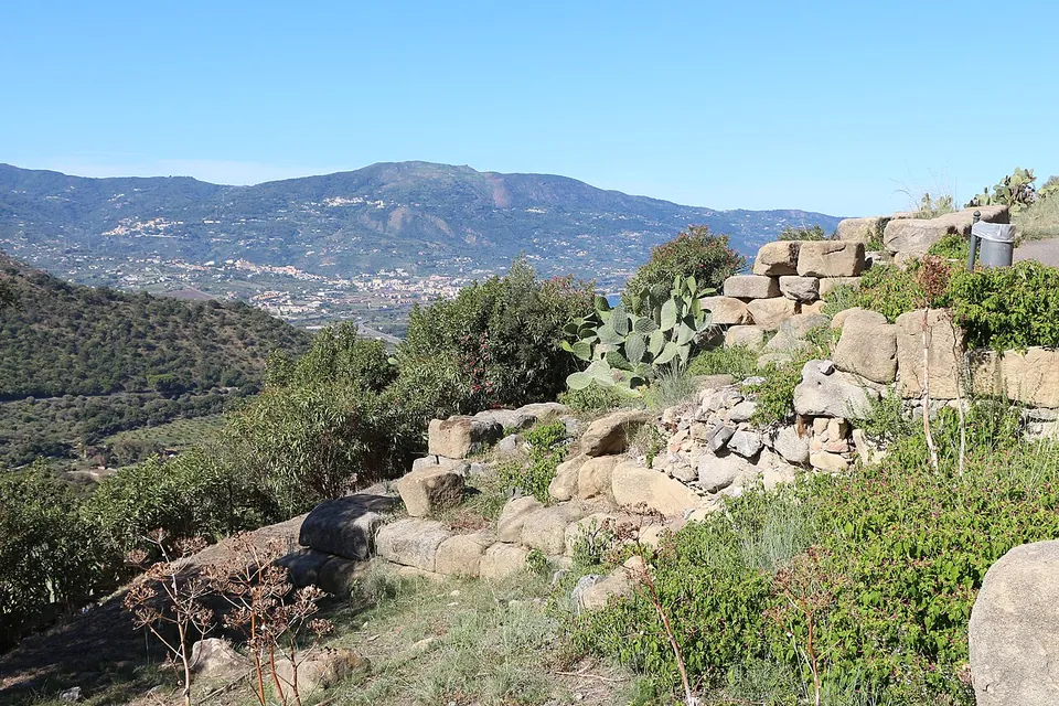 A line of large rough stone blocks running along a scrubby slope, with prickly pear growing beside it and a valley, a town and mountains inland in the distance.