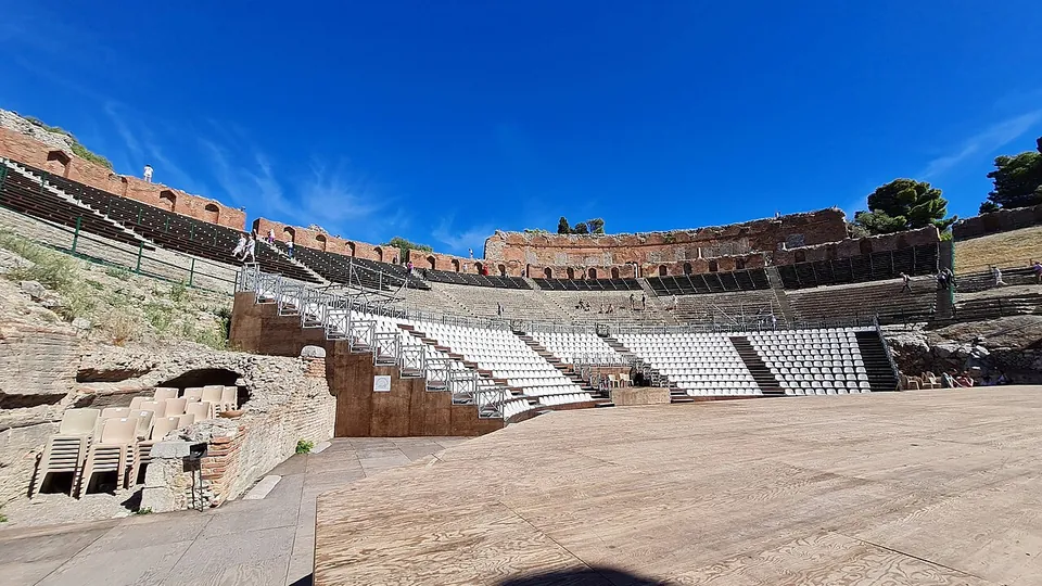 The seating bowl fitted with rows of modern white plastic seats on raised stands, seen from a wooden stage floor, with the brick upper wall of the theatre against a blue sky.
