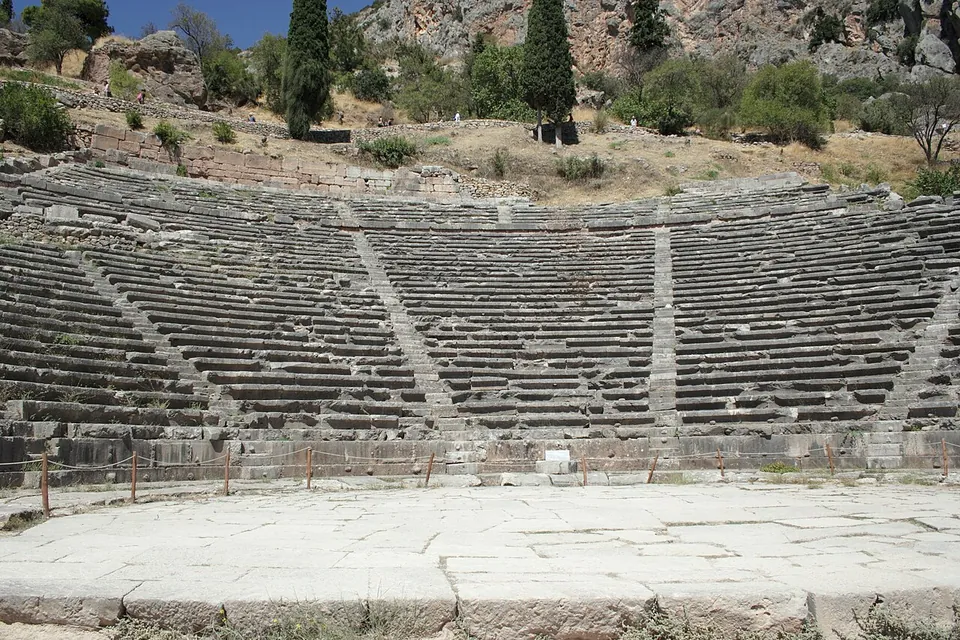 The curved tiers of a stone theatre rising up a hillside in sunlight, divided by stairways, seen from its paved floor, with cypresses and rocks above.