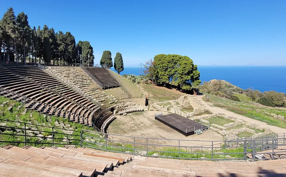 A semicircle of stone seating rows cut into a hillside, seen from the upper rows, with a low stage platform on the levelled floor below, umbrella pines and cypresses along the top, and the open sea beyond.