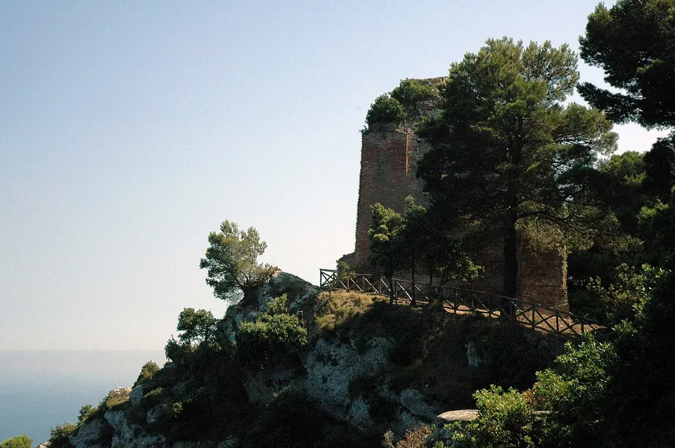 A square ruined tower of stone and brick rising among pines on a rocky outcrop, with a wooden-railed path running along the cliff below it and the sea far beneath.