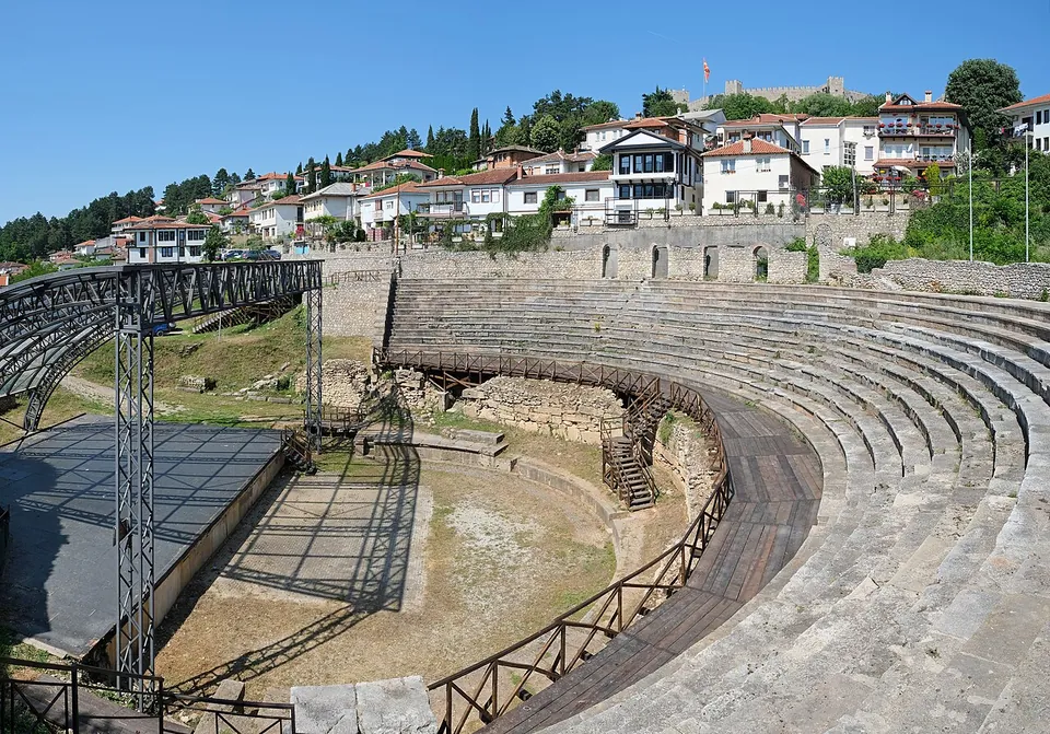 A semicircle of stone seating rows descending to a flat orchestra, with a metal stage structure on the left, whitewashed houses above and a fortress wall on the hilltop behind.