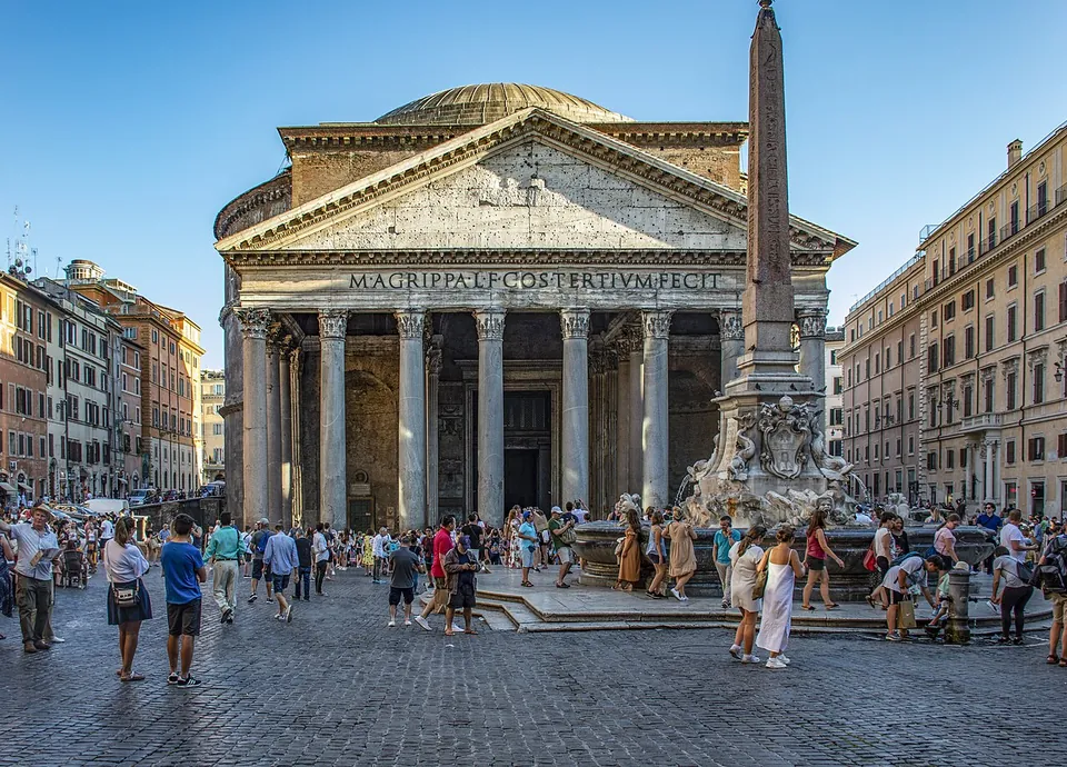 The Pantheon's portico across a busy square in strong sunlight, with a fountain topped by a tall obelisk in front of it and houses lining both sides of the square.