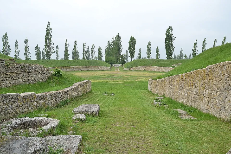 A long grassy arena between curving retaining walls of rough stone, with grass banks above the walls and a row of tall poplars along the far end.