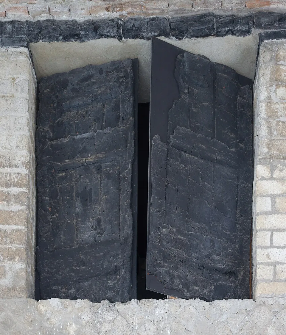 A pair of blackened, charred wooden shutters standing in a window opening framed by pale stone and brick.