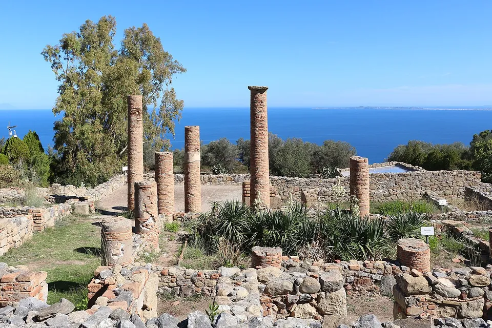 Several brick columns of different heights standing among low stone walls of an excavated house, with shrubs between them and the sea on the horizon.