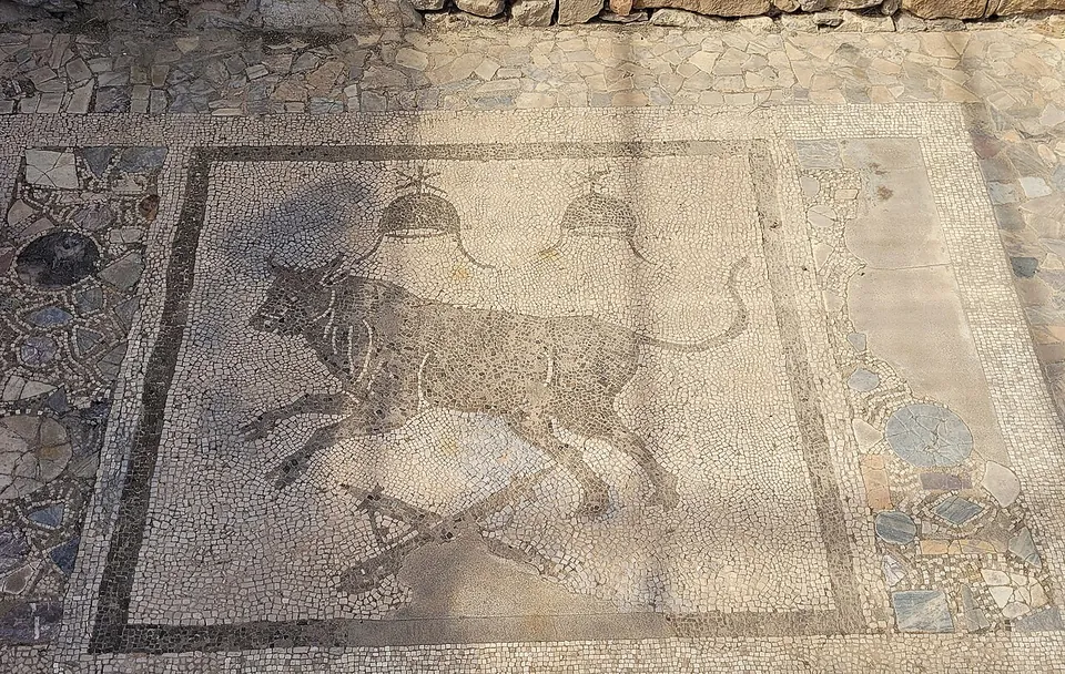 Close-up of a black-and-white mosaic panel showing a running bull, with two conical caps topped by stars above it, framed by a border of coloured stone pieces.