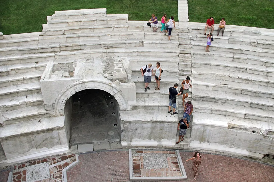 Looking down on the white marble tiers of the stadium's curved end, with visitors sitting and climbing on them and a dark arched passage leading in under the seats.