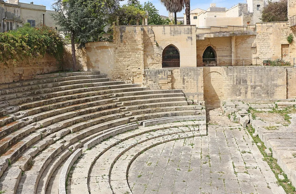 Curving rows of pale stone seating steps run round a paved semicircular floor, closed off behind by the plain stone walls and arched windows of later buildings, with trees and palms above.