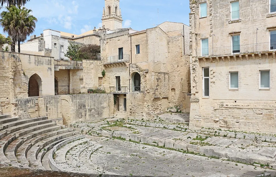 The lower seating rows and the stone floor of the theatre in the foreground, hemmed in by tall pale stone houses and garden walls, with a bell tower rising behind them.