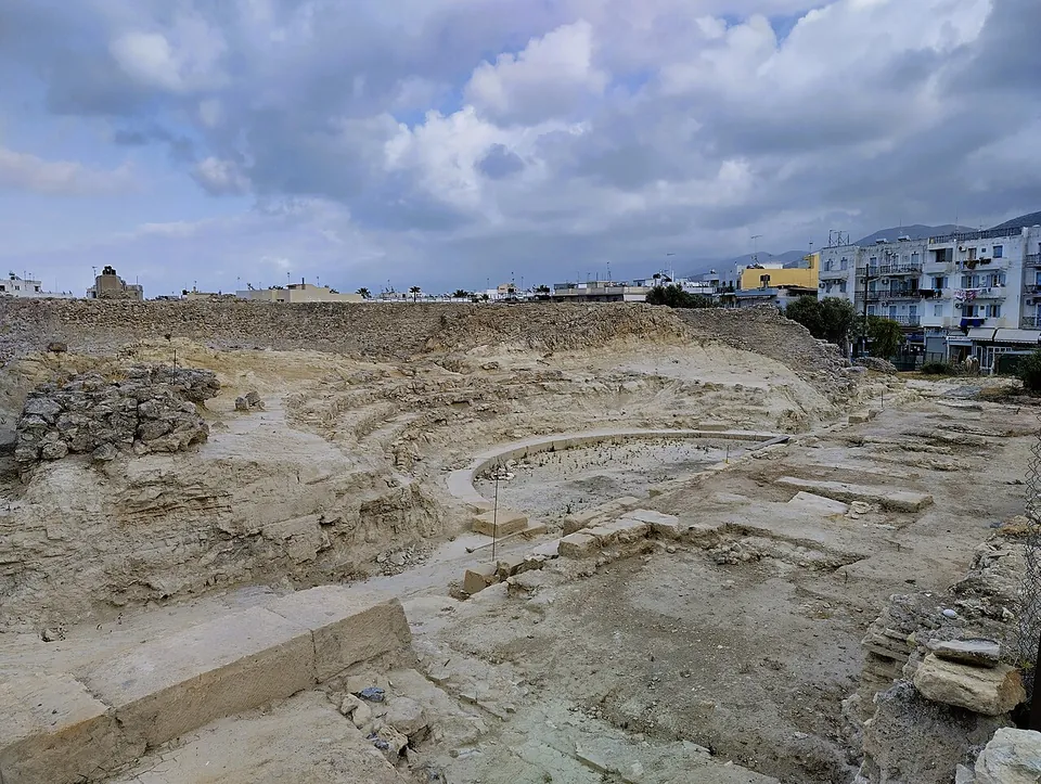 The excavated seating of a theatre, low tiers of pale stone curving round a semicircular orchestra, with the stone foundations of the stage in front and apartment blocks behind under a cloudy sky.